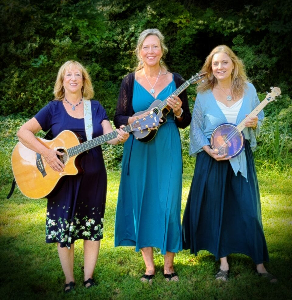 Suzanne Brindamour, Michelle Lockey, and Michelle Murray standing in a bright green field holding a guitar and ukuleles
